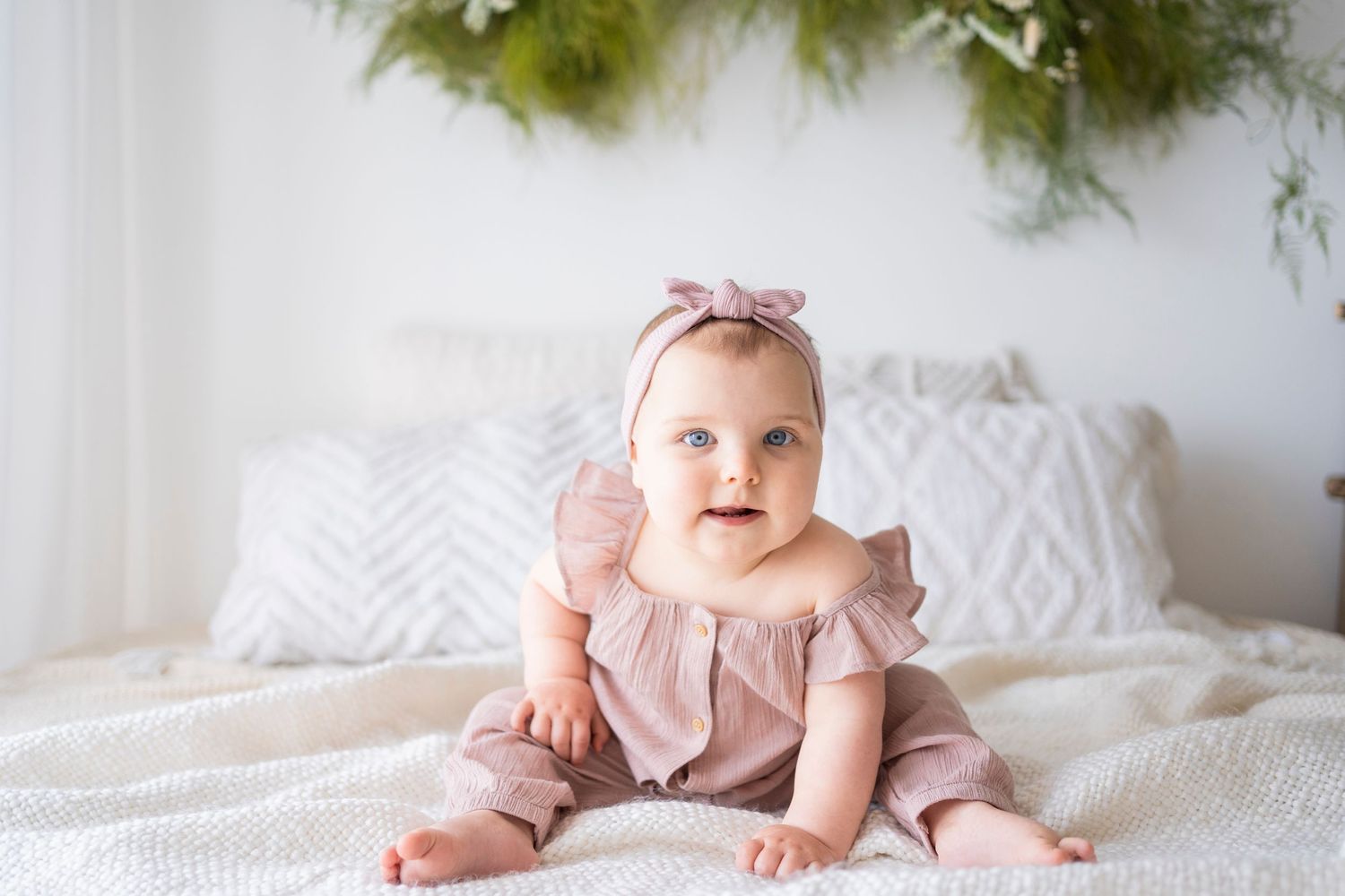Adorable baby girl in a dusty rose ruffle outfit and bow headband sitting on a white bed.