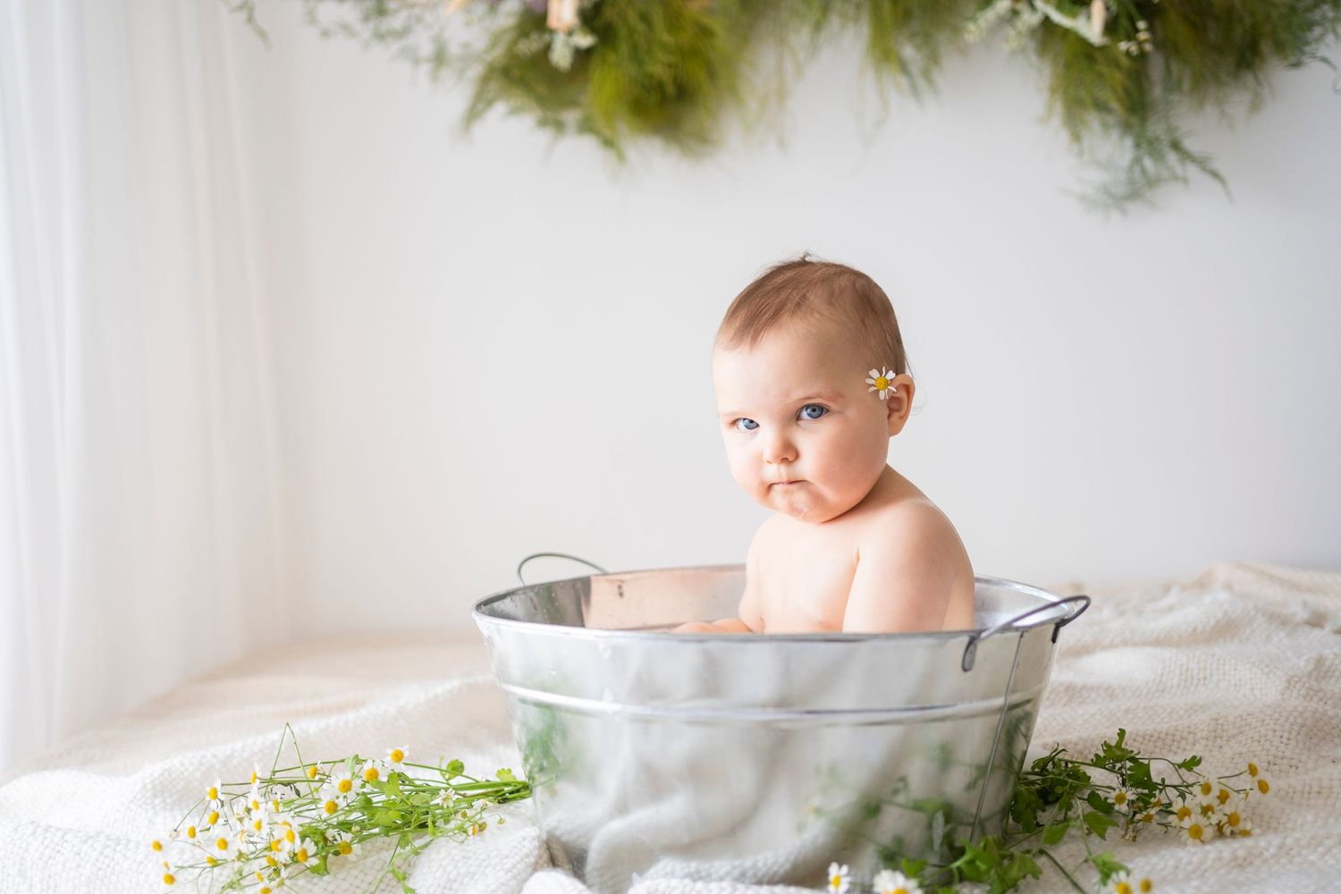 Baby with serious expression sitting in galvanized tub with green floral garland backdrop.