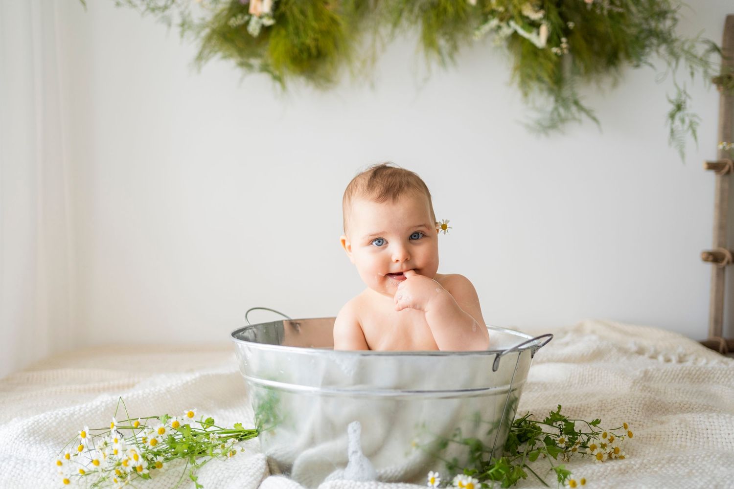 Cheerful baby smiling in vintage metal tub surrounded by wildflowers and lush green garland.
