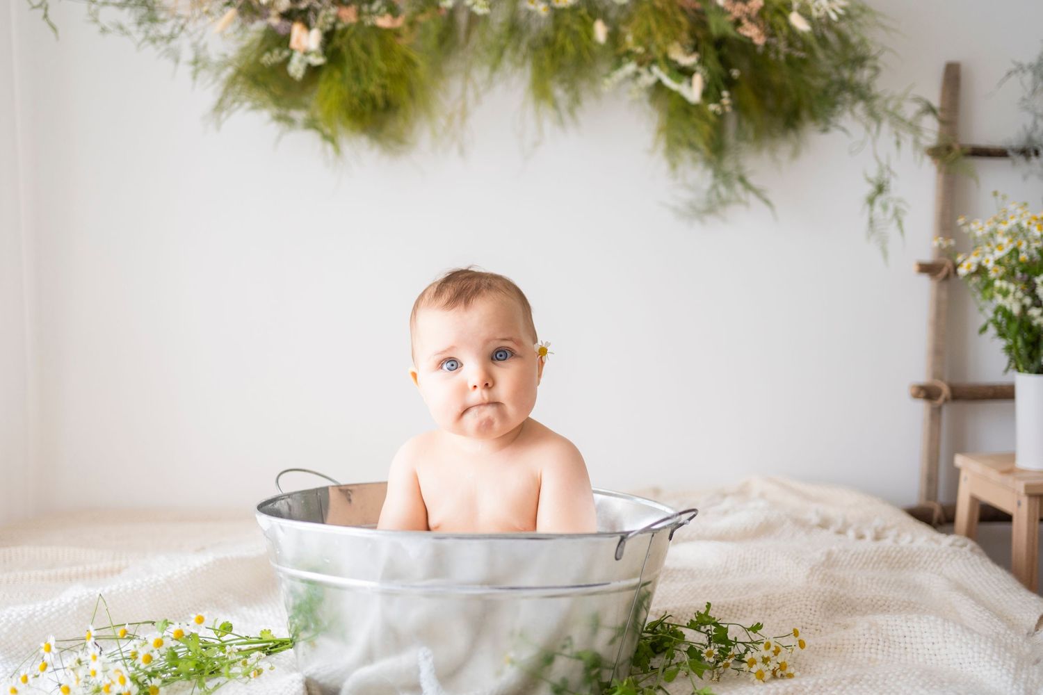 Cute baby sitting in a vintage metal tub surrounded by wildflowers and greenery in a bright studio.