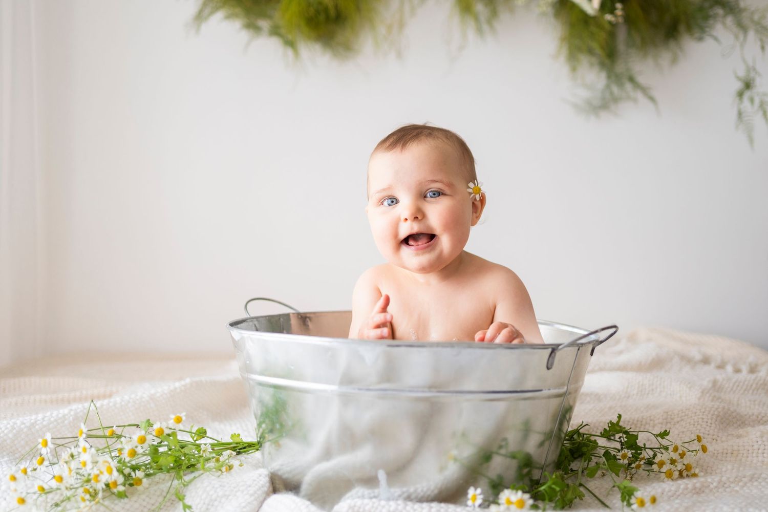 Happy smiling baby sitting in a galvanized metal tub with chamomile flowers in a bright white studio.