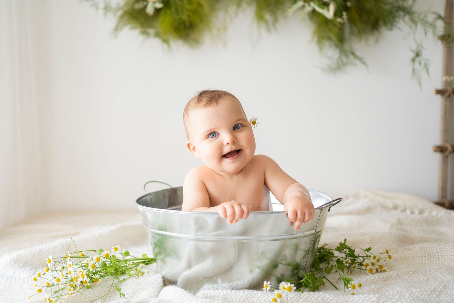 Joyful baby smiling in a vintage galvanized tub with daisy flowers and lush green garland decor.