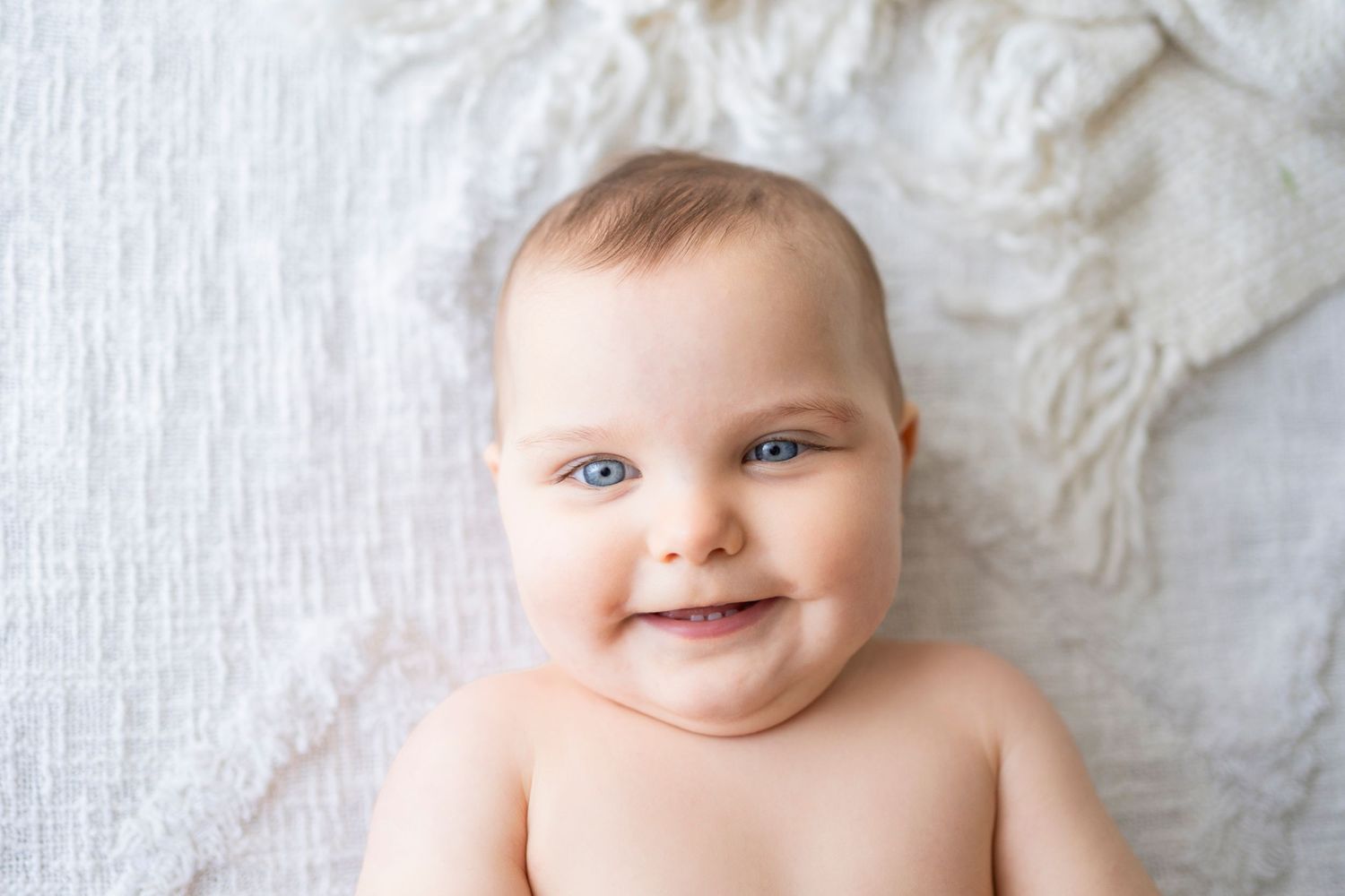 Close-up portrait of a happy blue-eyed baby smiling while lying on a soft white knit blanket.