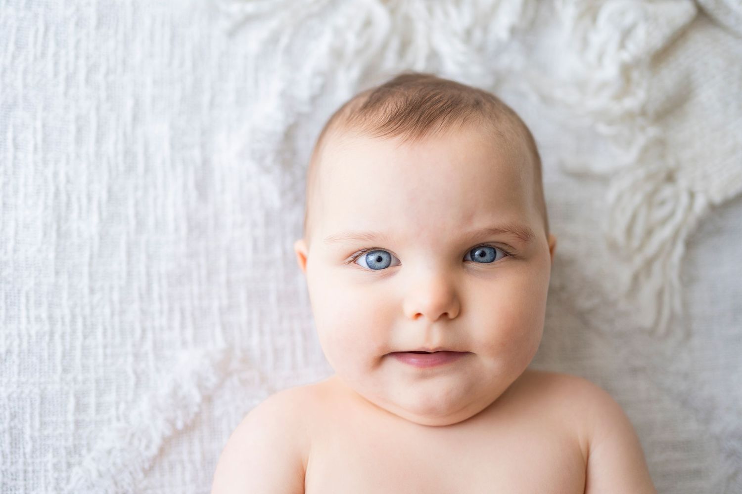 Adorable blue-eyed baby with a gentle smile lying on a soft white textured blanket in studio.