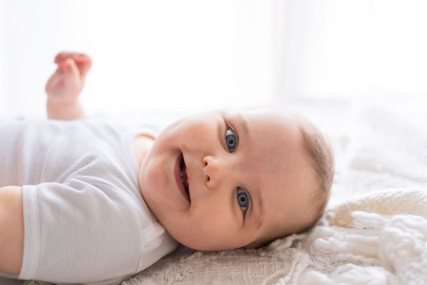 Sweet smiling baby in a white onesie lying on a cozy white blanket with arm raised playfully.
