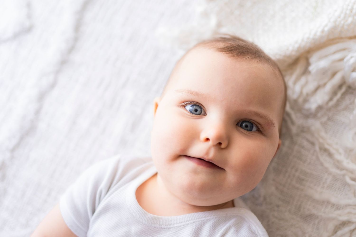 Cute blue-eyed baby in a white onesie looking up with a gentle smile on a soft white blanket.