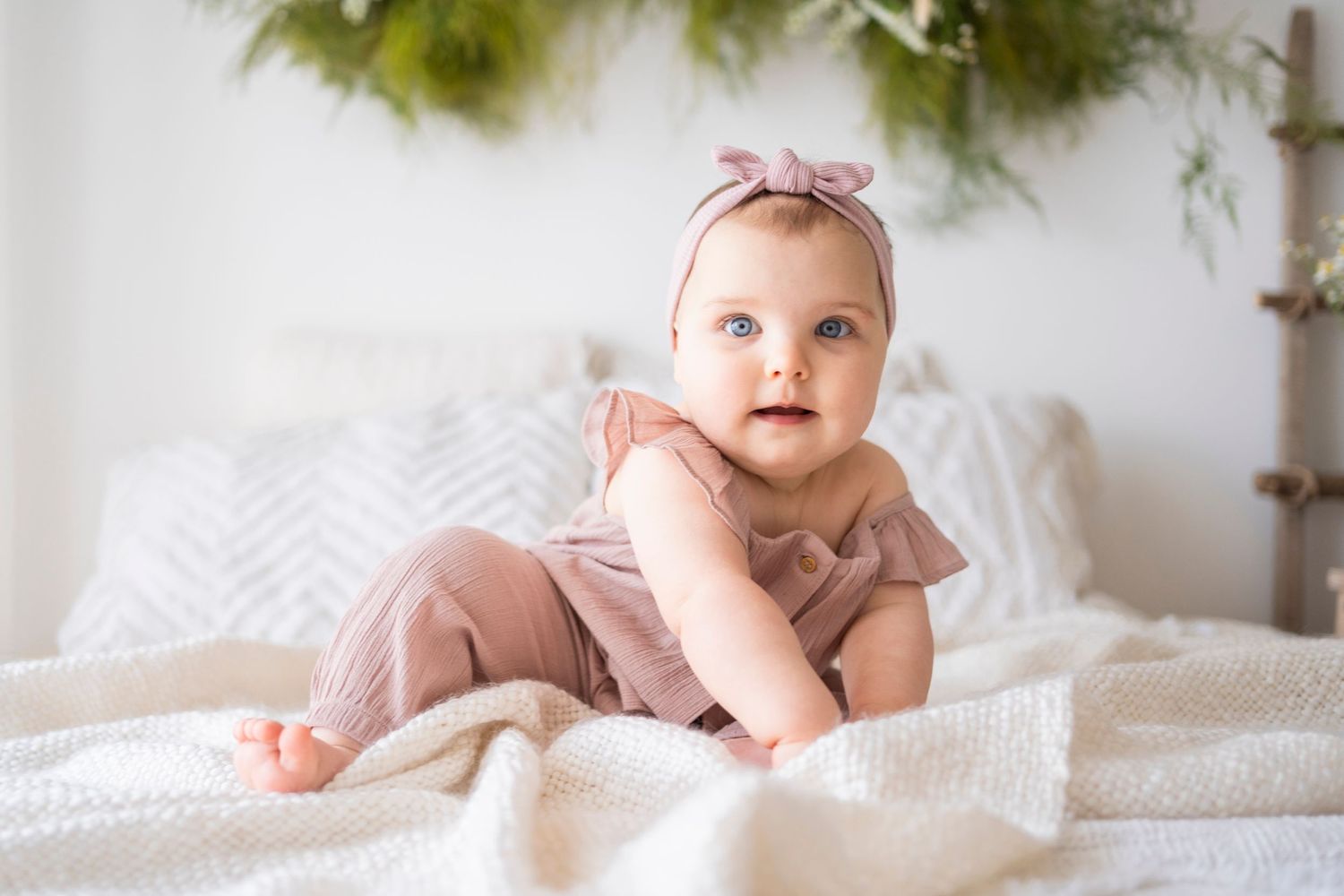 Adorable baby girl in pink romper and bow headband doing tummy time on white bed with greenery backdrop.