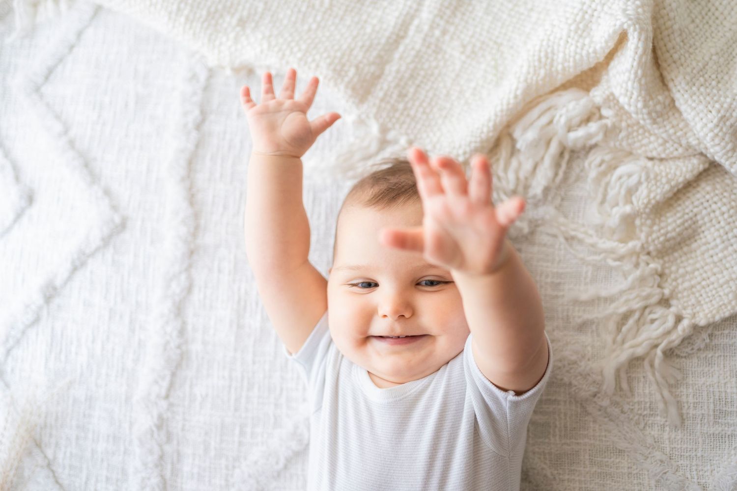 Happy baby in white onesie lying on blanket with both hands raised and a big cheerful smile.