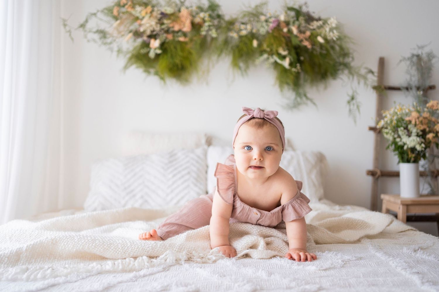 Sweet baby girl in dusty rose outfit crawling on white bed with floral greenery wall decoration.