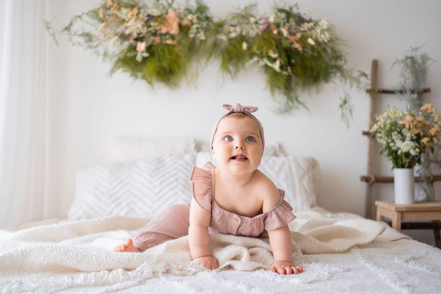 Curious baby girl in pink ruffle romper looking up while doing tummy time on white bed with flowers.