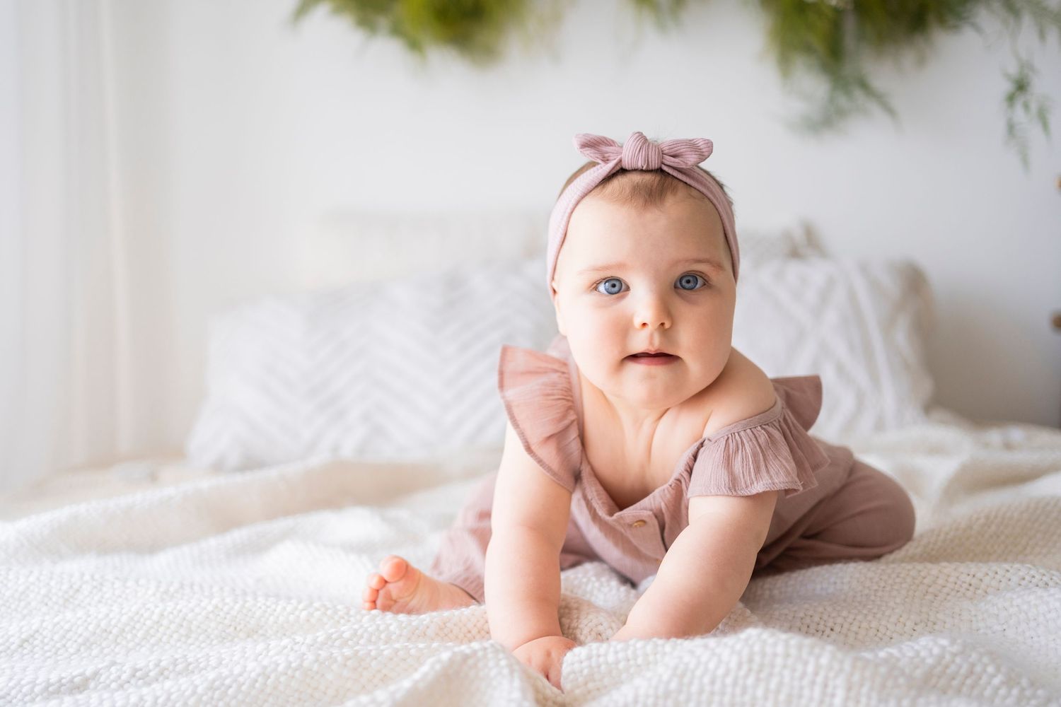 Adorable baby girl in pink ruffle outfit and bow headband crawling on white bed.