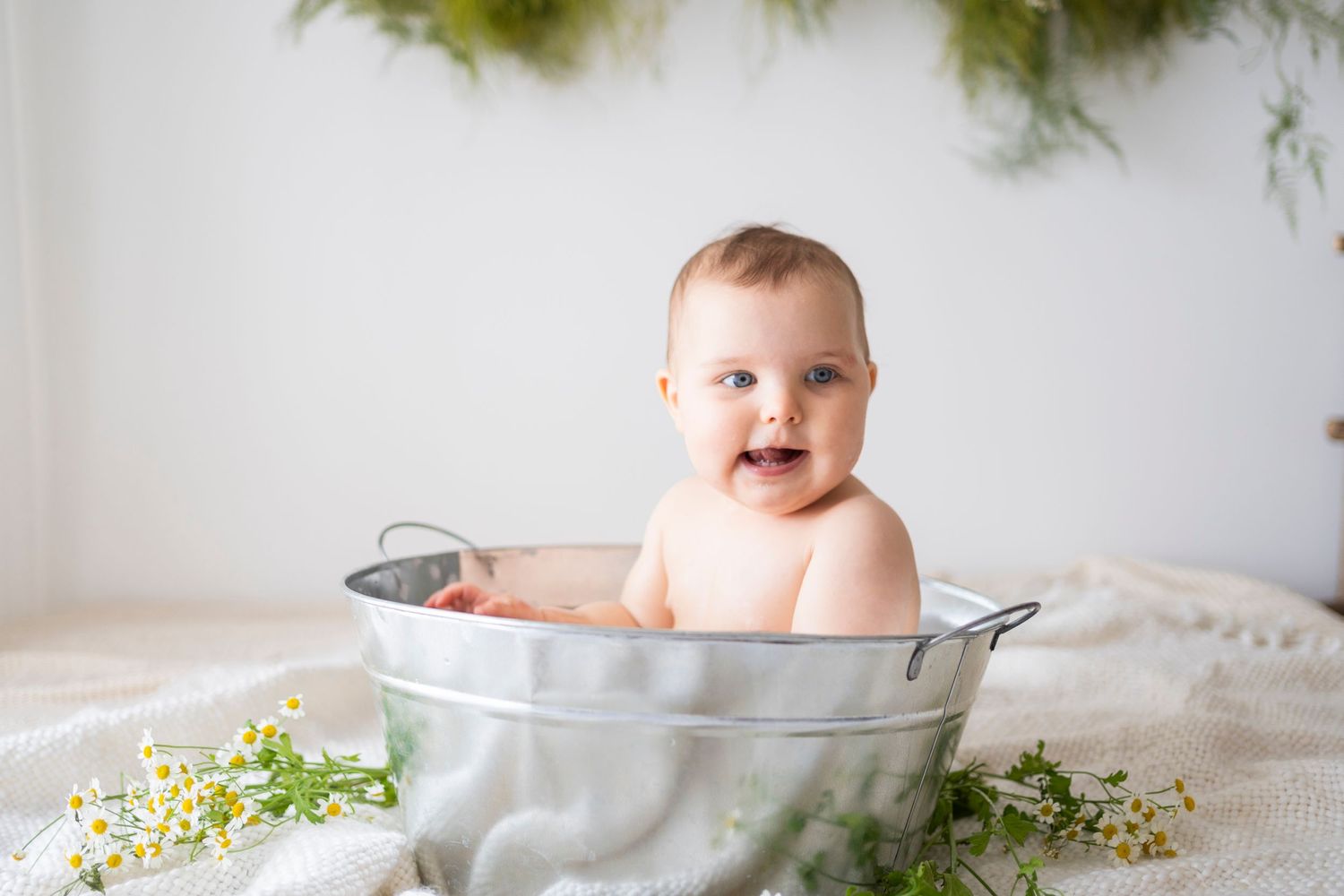 Happy laughing baby with blue eyes sitting in silver metal tub with floral decor.