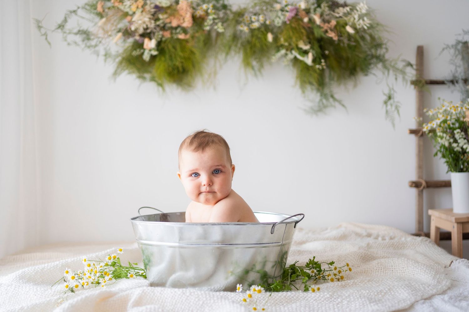 Cute baby sitting in vintage galvanized tub with milk bath surrounded by chamomile flowers.