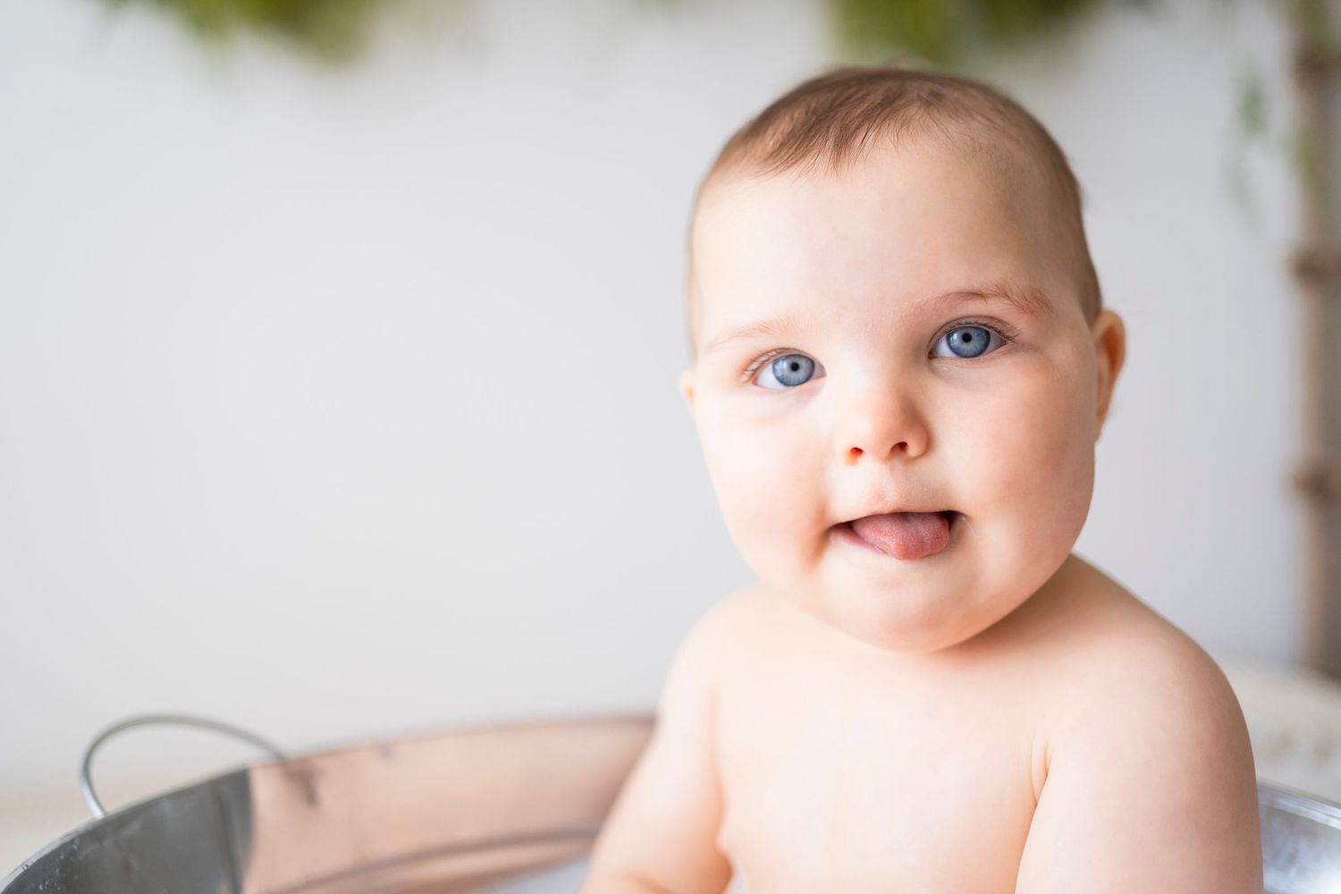 Close-up of smiling blue-eyed baby in metal tub with soft natural light background.