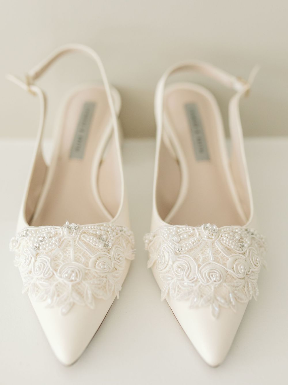 Elegant white bridal shoes with delicate lace detailing displayed on light background.