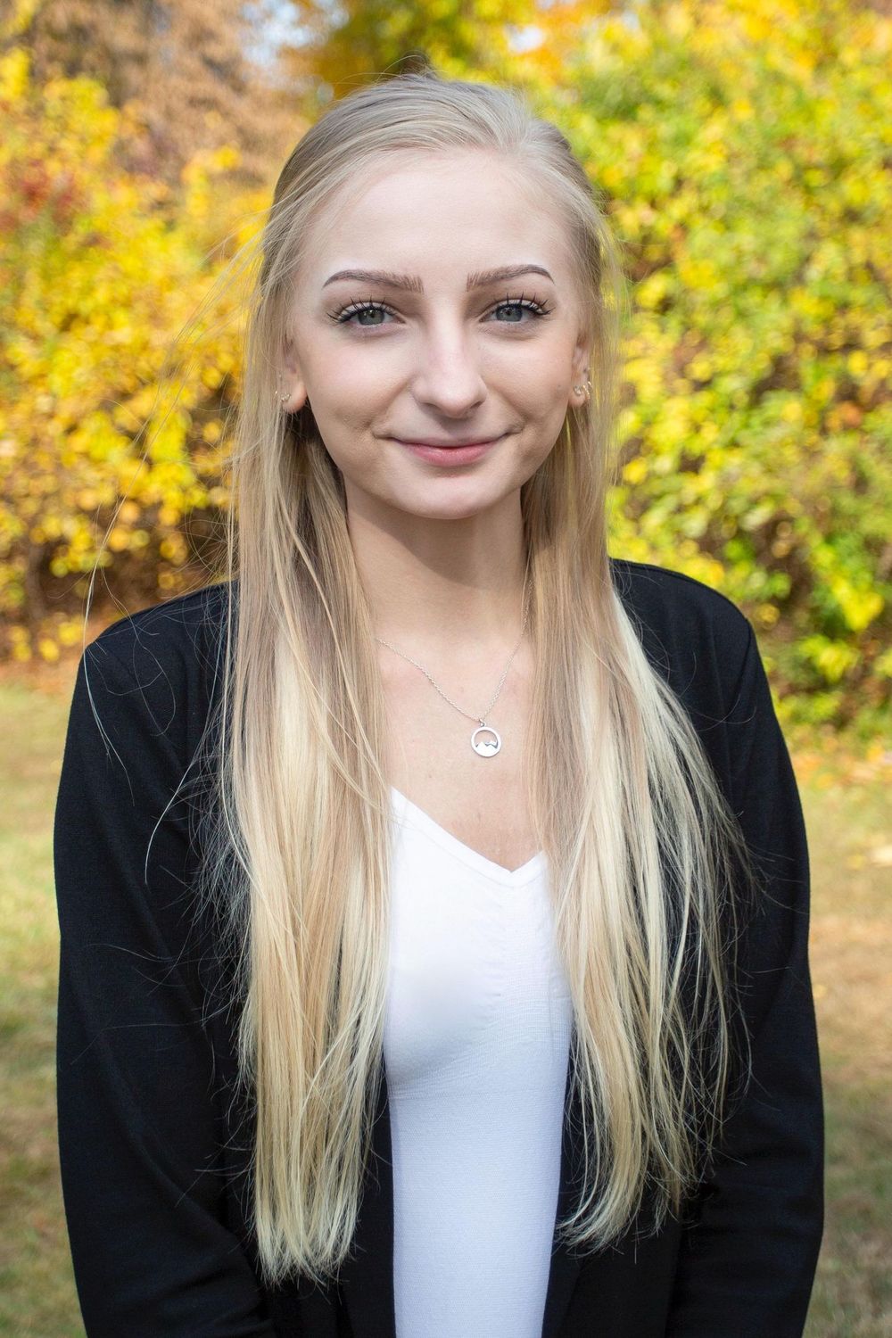 Portrait with long blonde hair wearing black cardigan and white top against autumn foliage.