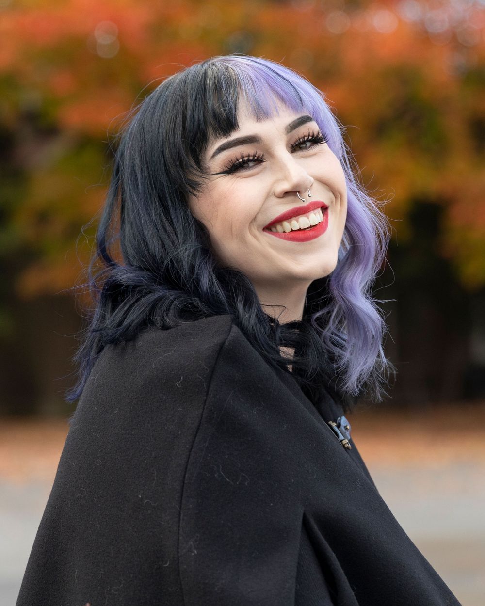 Stylish portrait with black and purple hair wearing dark clothing against autumn foliage.