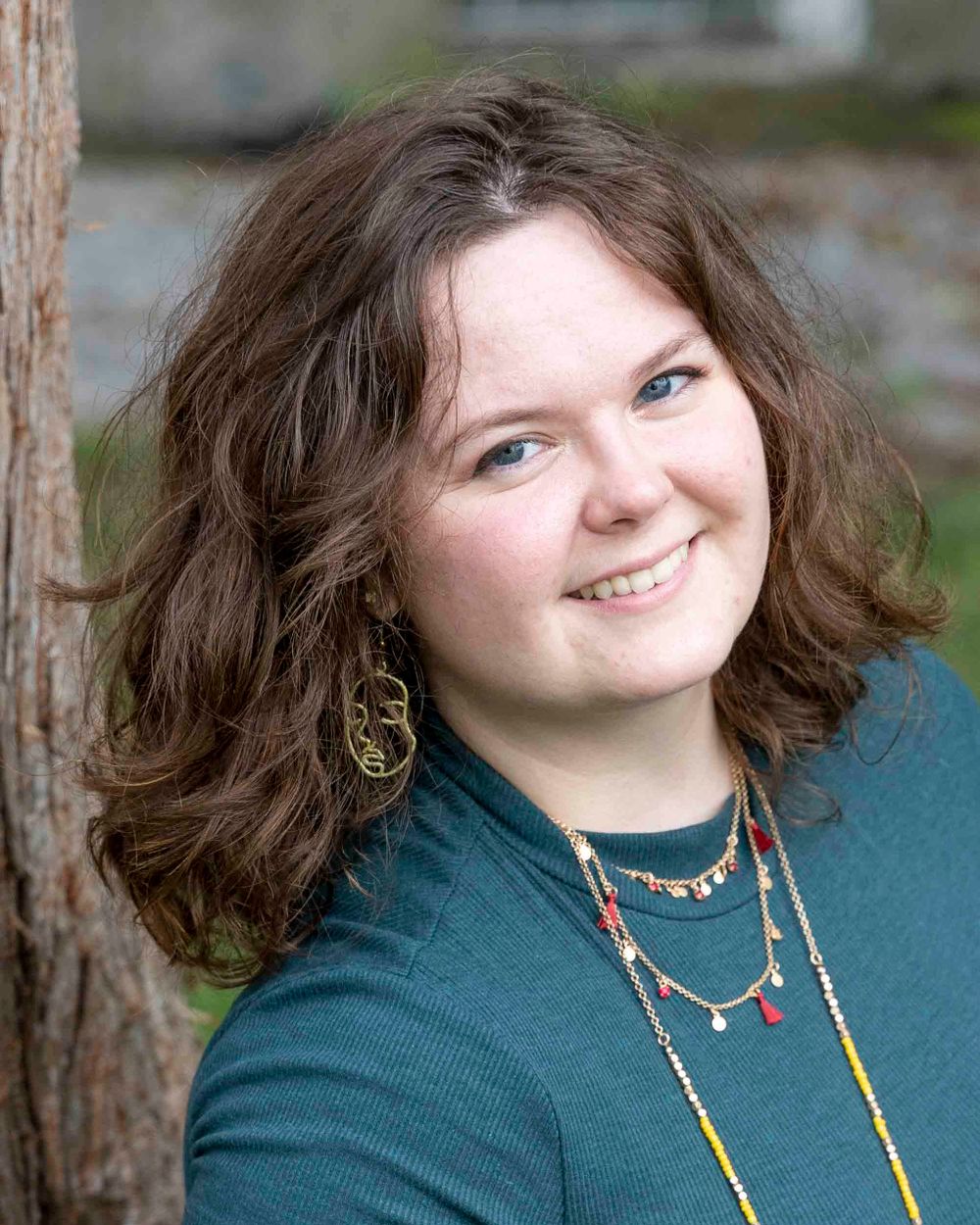 Portrait of a person wearing a teal top and layered necklaces with curly brown hair leaning against a tree trunk.