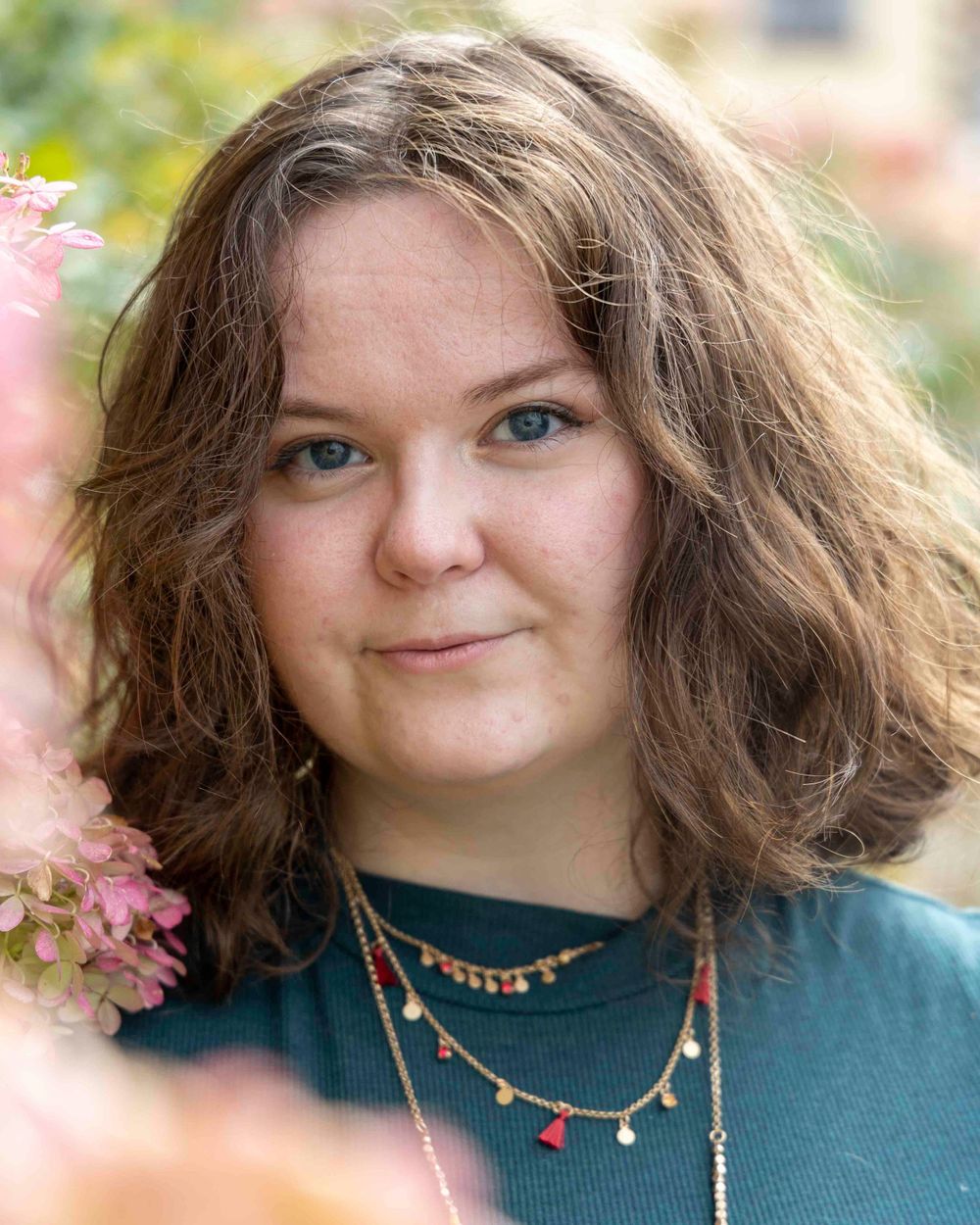 Close up portrait showcasing wavy brown hair and layered necklaces against a soft floral background.