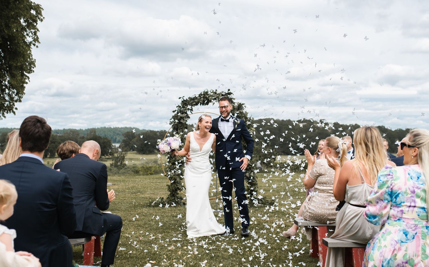 Wedding ceremony guests throw confetti at a celebration under a floral arch in an outdoor setting.