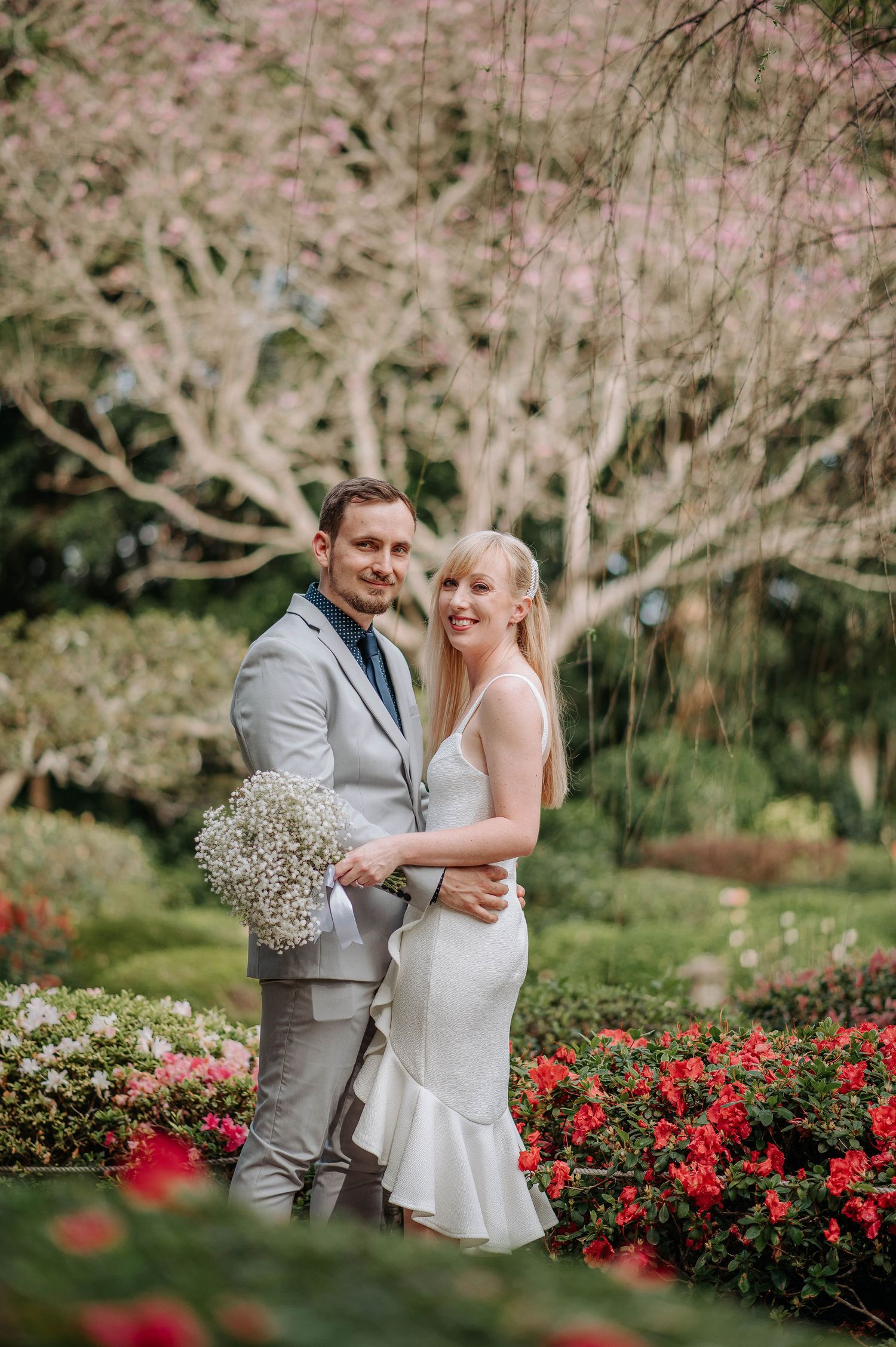Wedding couple poses with bouquet in garden with blooming red flowers and cherry blossoms.