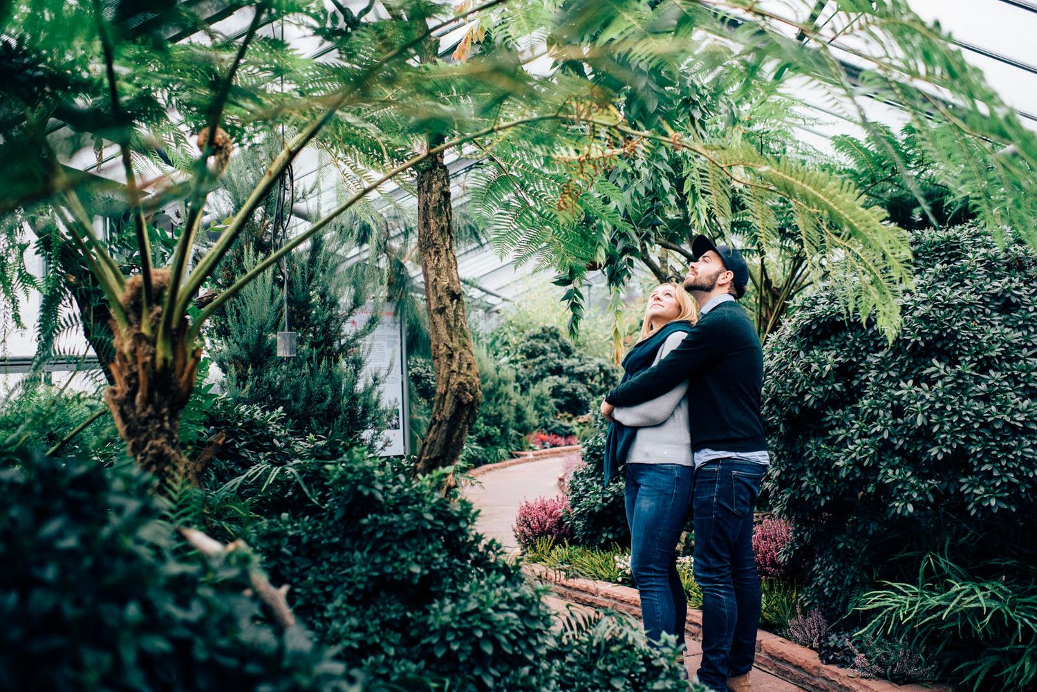 A couple embracing in a lush greenhouse filled with tropical plants and natural light.