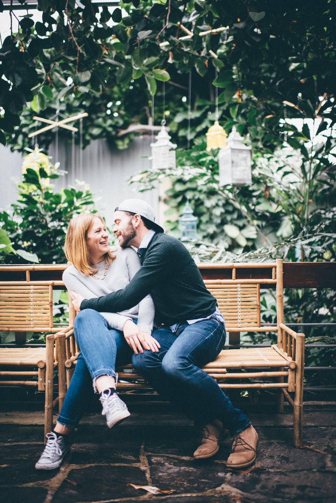 A couple sits close together on a wooden bench surrounded by greenery and hanging lanterns in a garden setting.