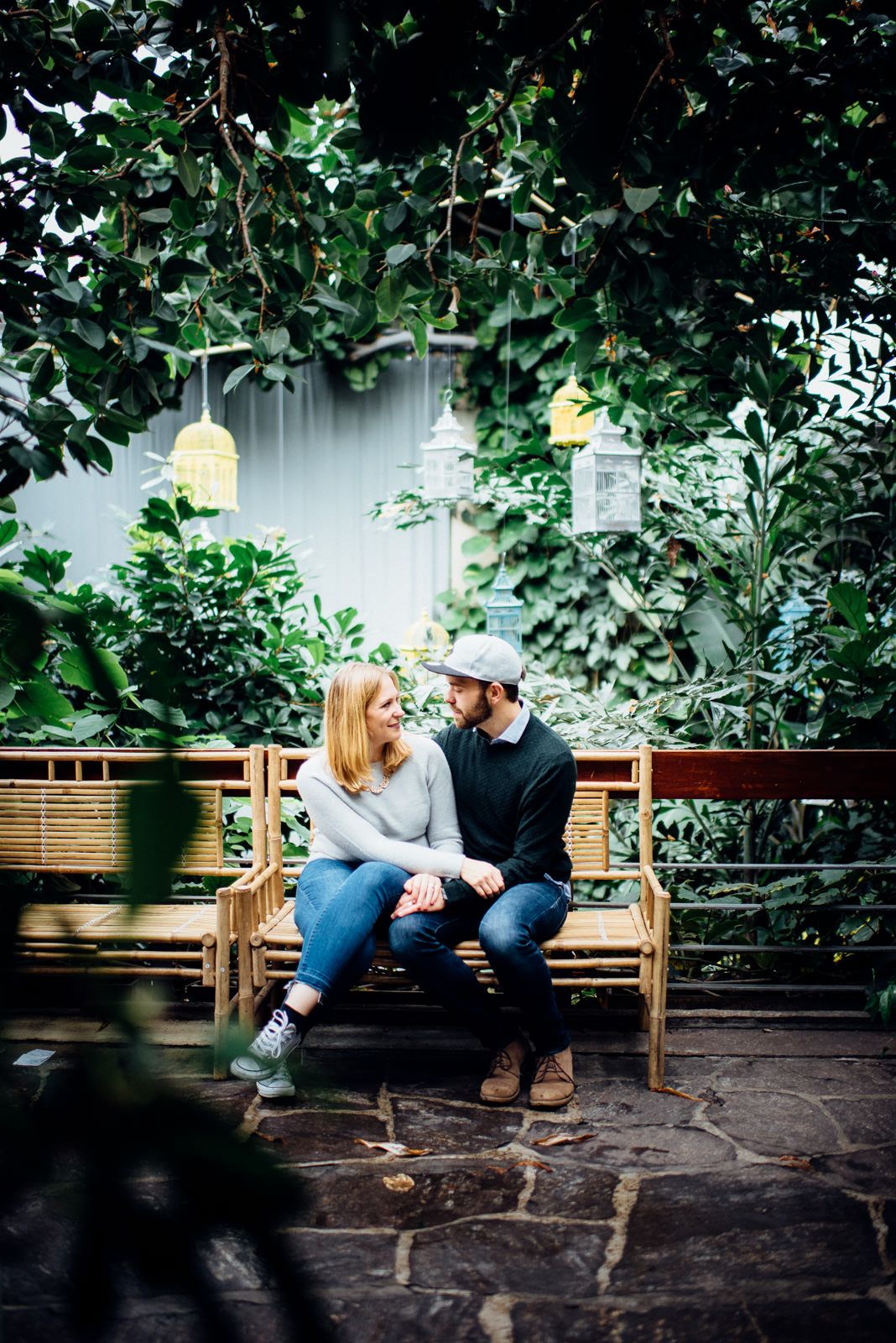 Romantic garden scene with a couple on a wooden bench under hanging lanterns and lush foliage.