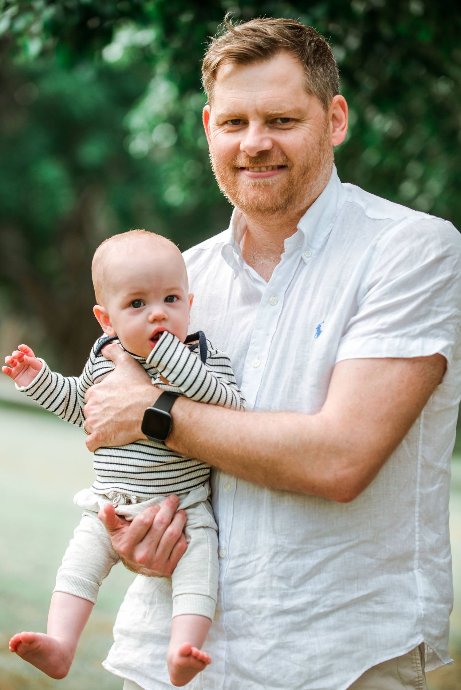 Father in white shirt holding baby in striped outfit during outdoor family portrait session.