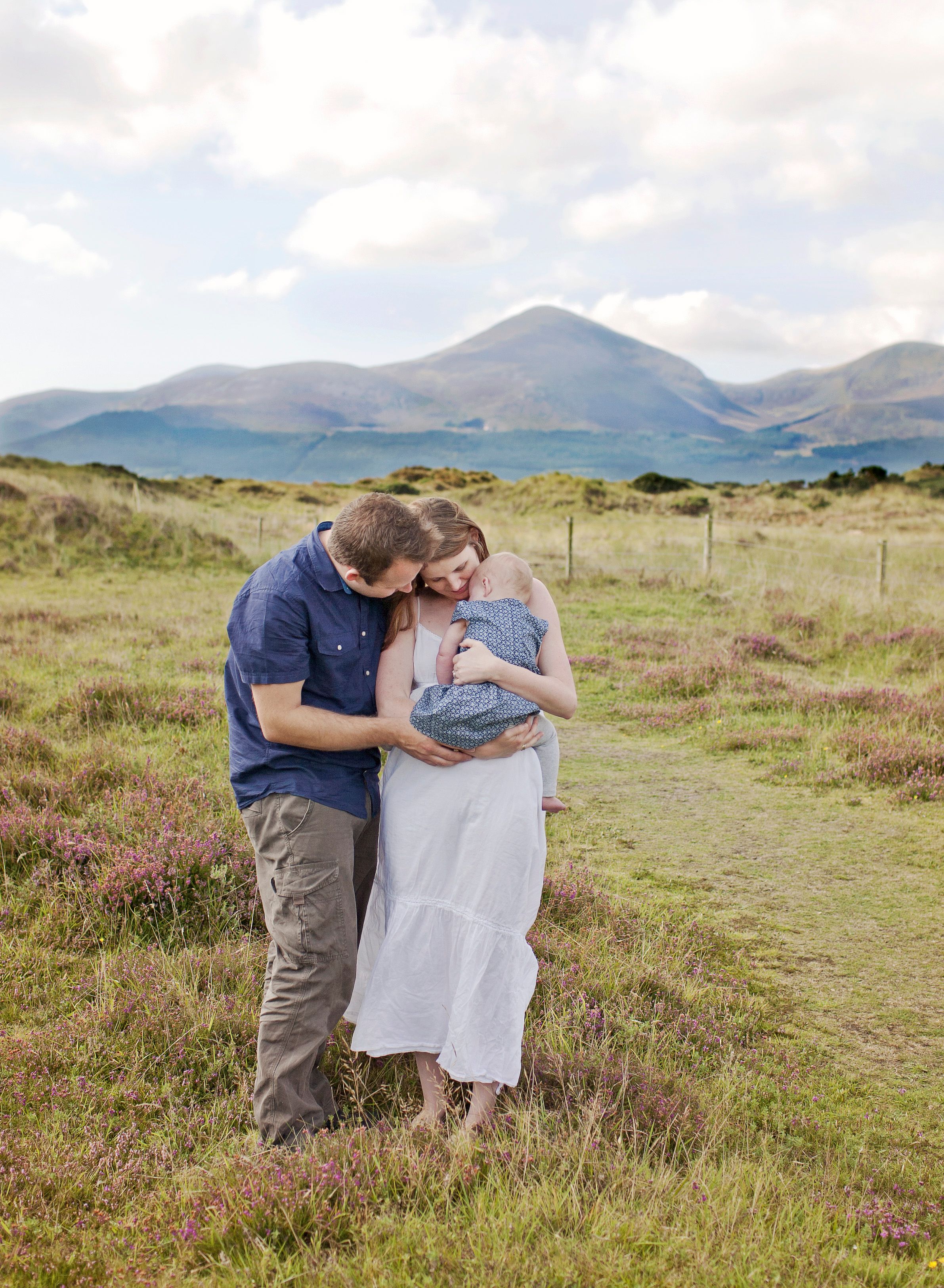 Couple embraces on scenic hilltop path with mountains and water in distance.