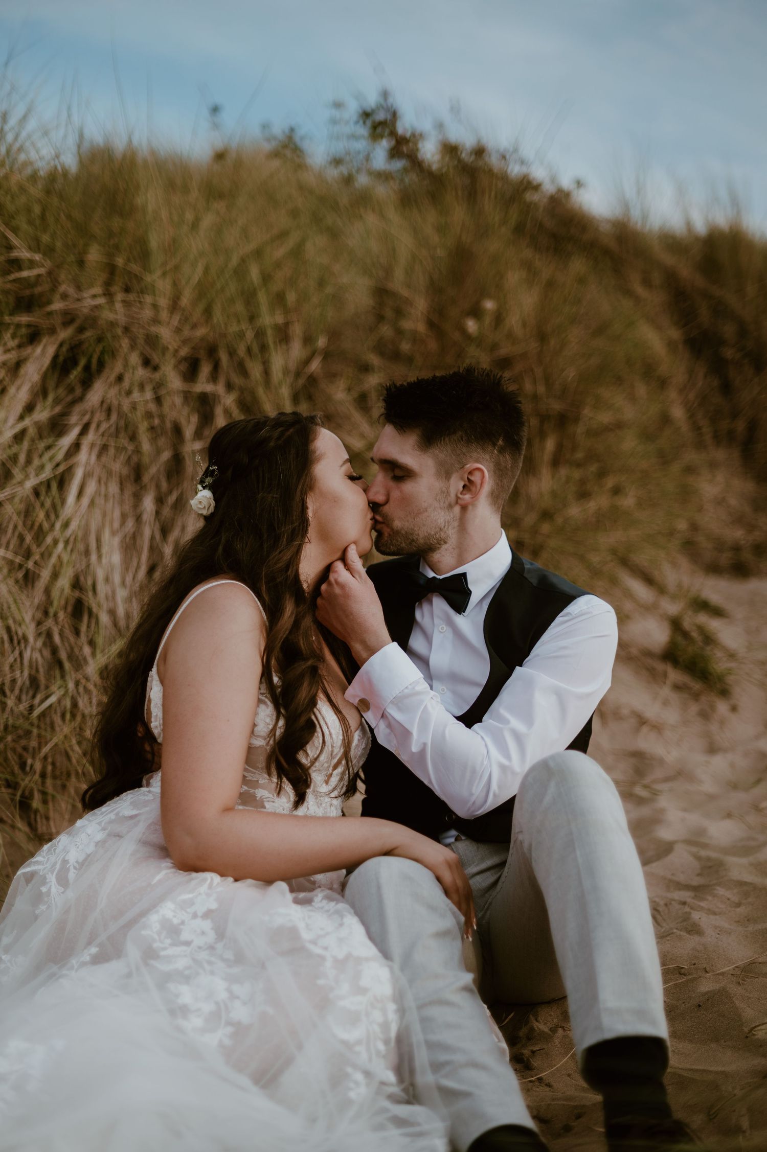 A romantic wedding couple shares an intimate moment while sitting together on sandy dunes at golden hour.