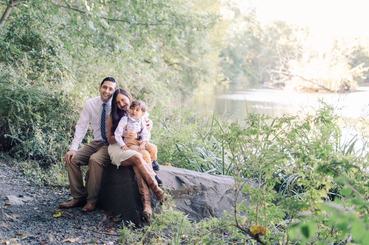 Family of three sitting on a rock by a river surrounded by lush greenery.