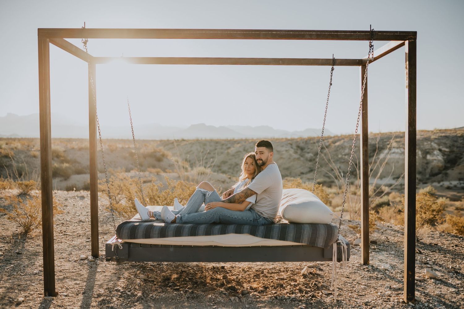 Couple relaxing on suspended outdoor bed frame in desert landscape at sunset.
