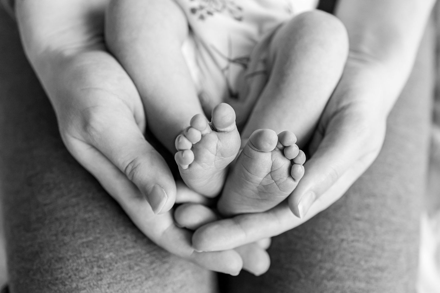 Close up of an adults hands, with newborn feet cupped in them, black and white, adult legs underneath the cupped hands