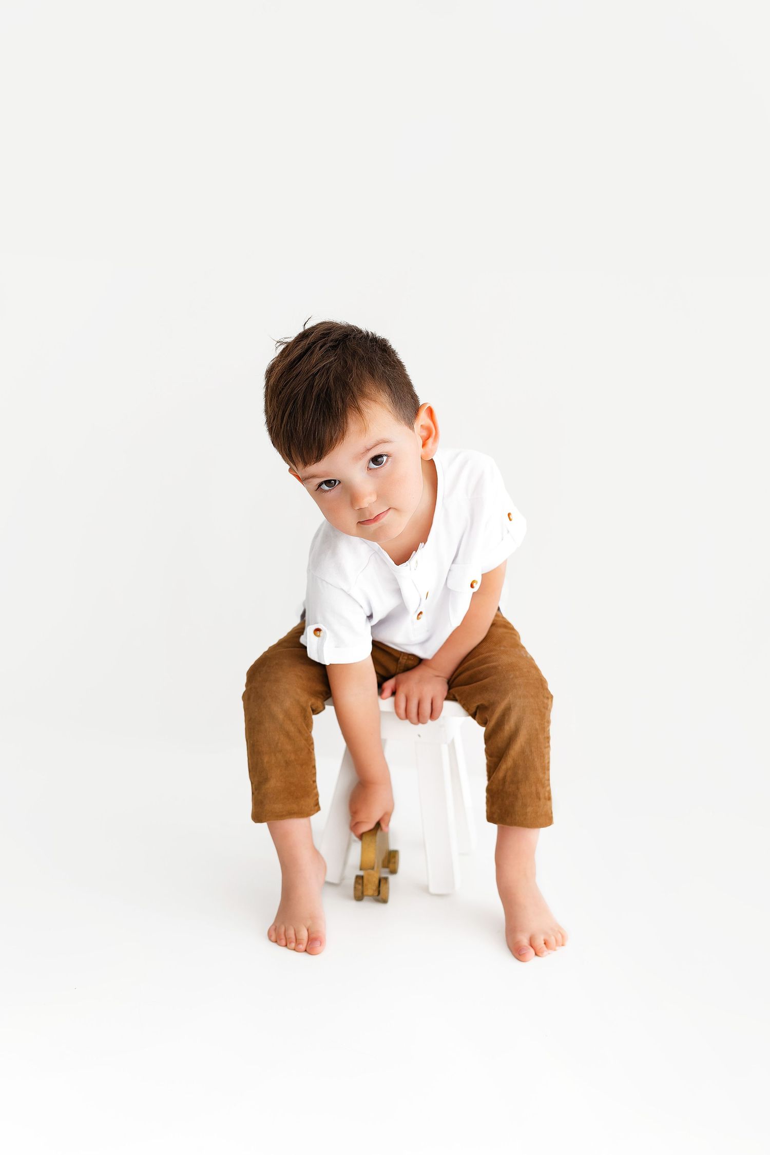 Young child in brown pants and white shirt sitting casually on white stool against white background.