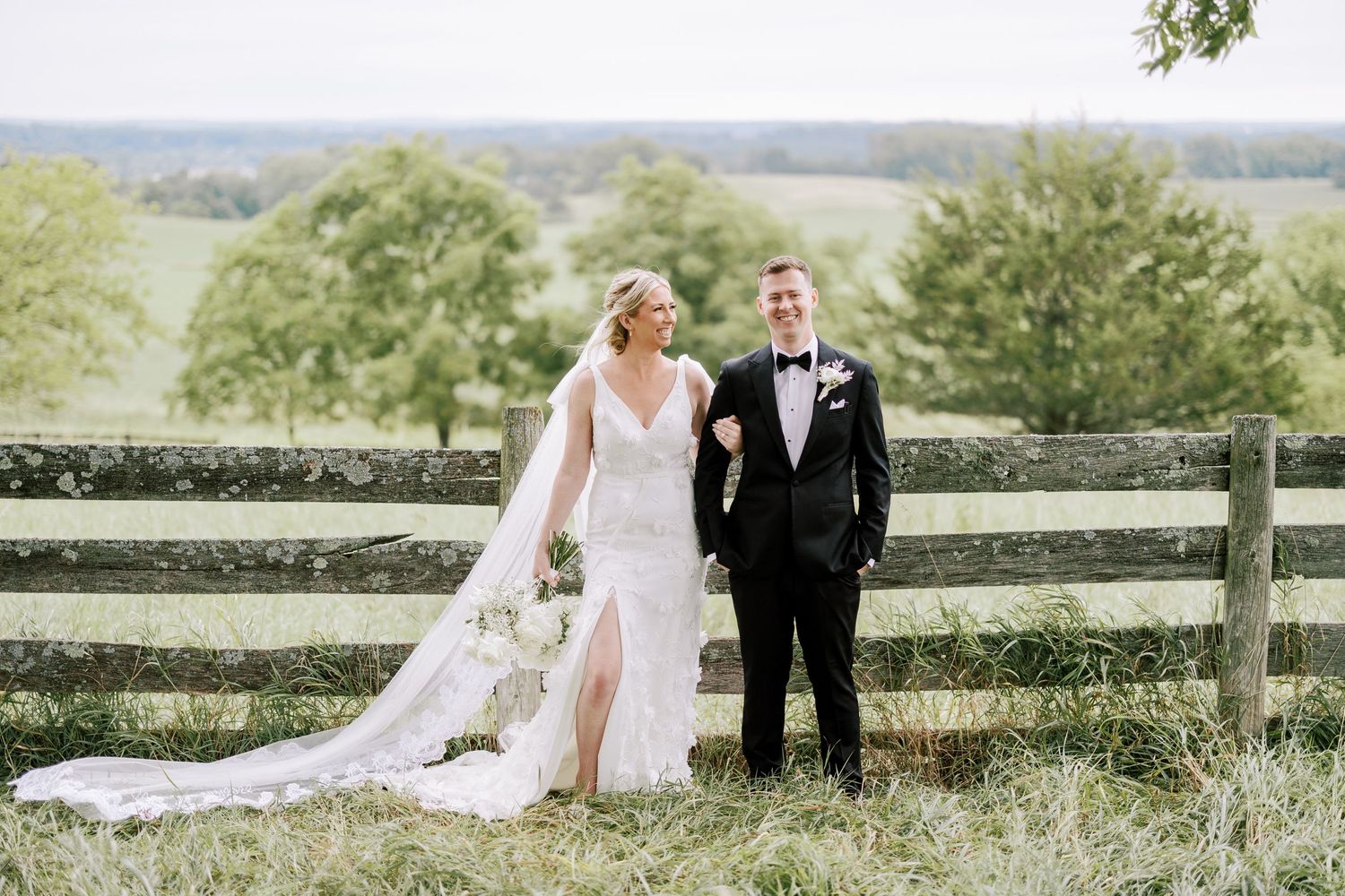 Newlyweds share intimate moment on rustic farm with wooden fence and rolling hills.