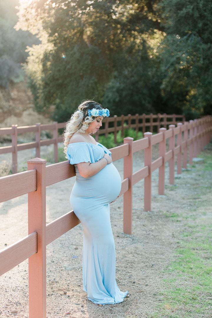 Maternity photos of woman in light blue dress posing by wooden fence at sunset.