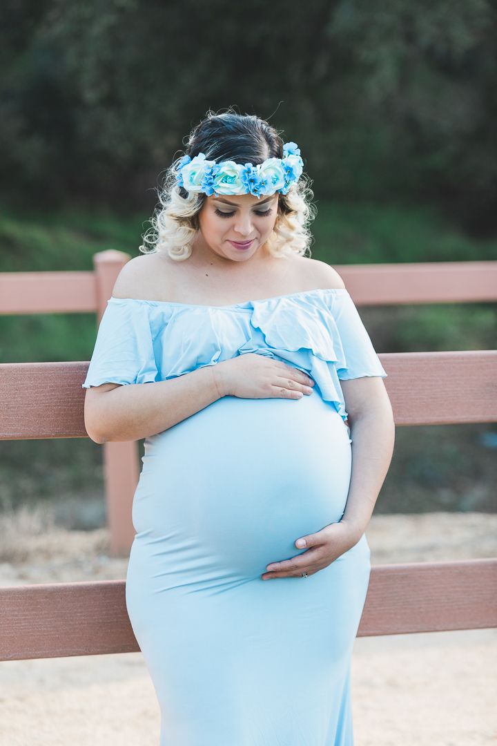 Maternity photo session outdoors with woman in light blue off-shoulder dress and floral crown against natural background.