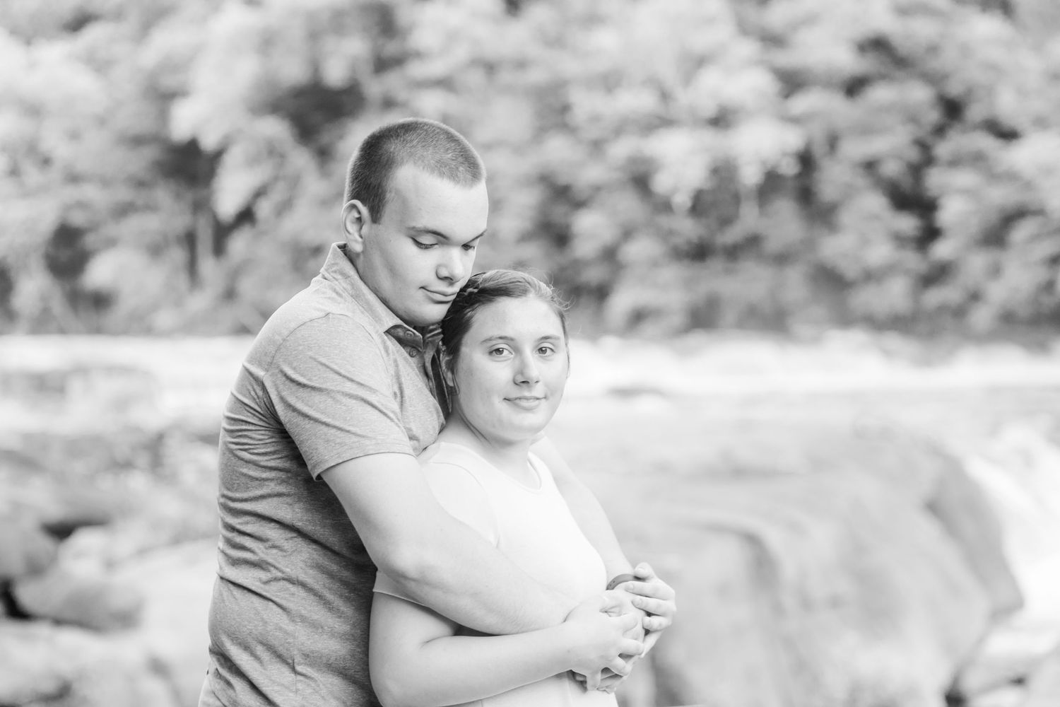A black and white portrait of two people embracing outdoors with natural scenery in the background.