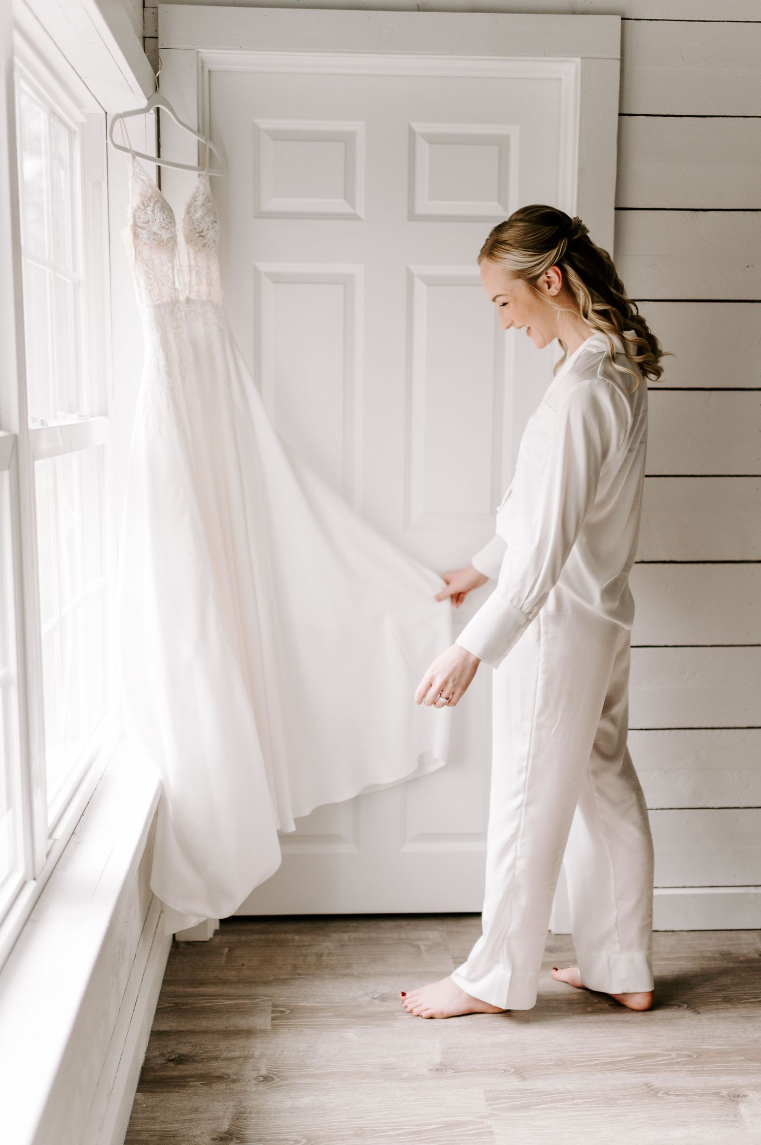 Person in white attire admires wedding dress hanging in bright room with shiplap walls.