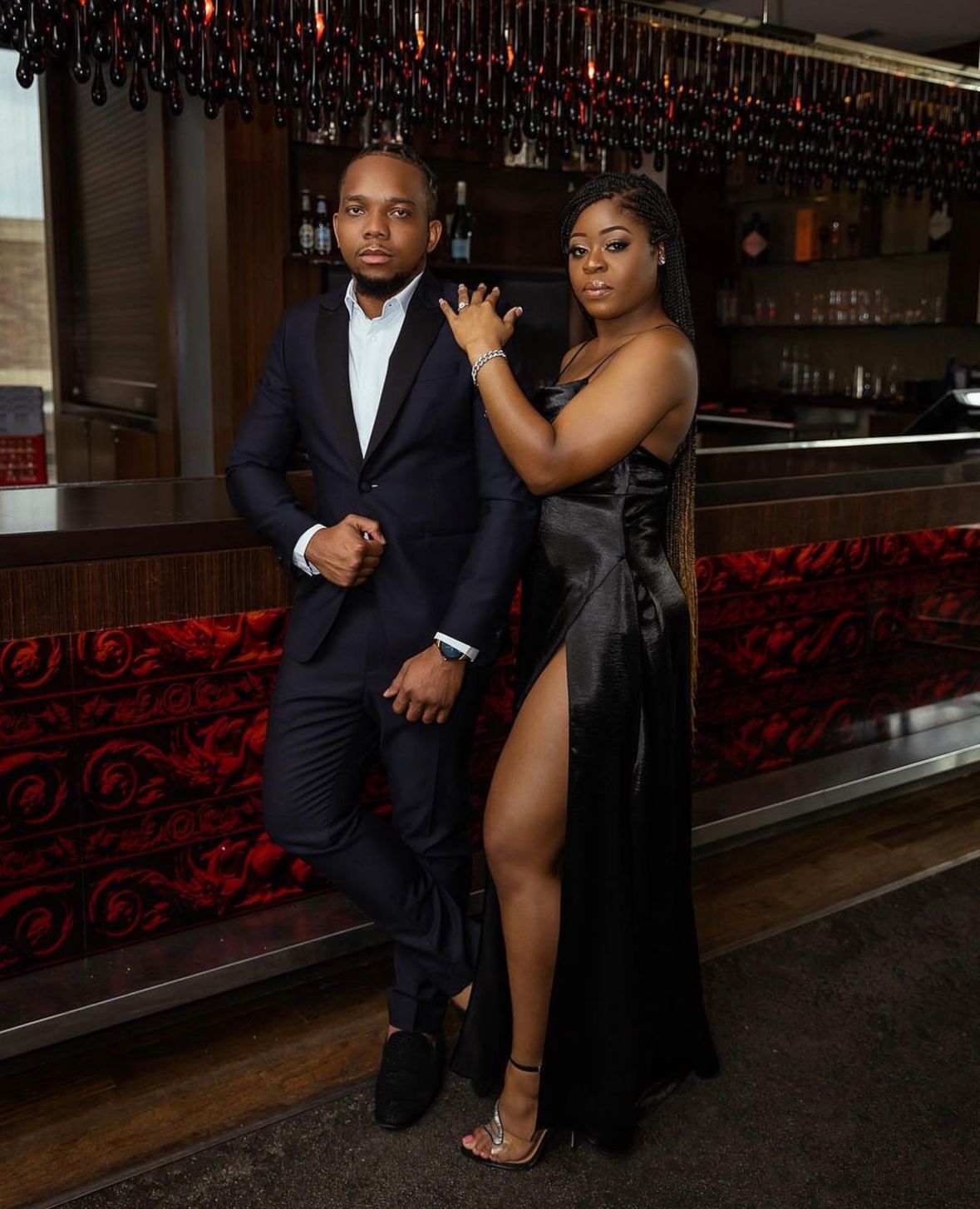 Couple in formal attire poses in front of a bar with ornate red decor.