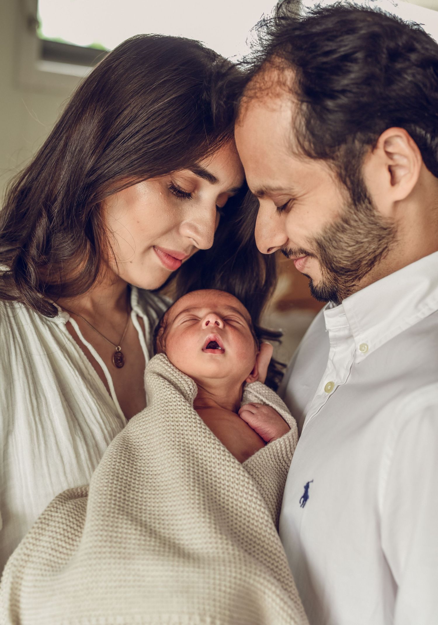 Intimate indoor portrait of new parents with newborn wrapped in cream blanket.