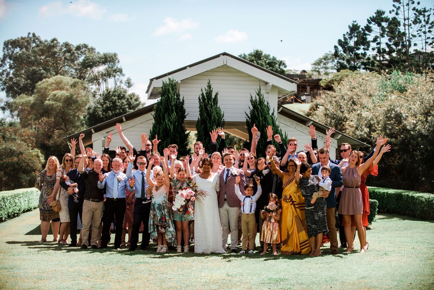 Large group wedding photo with guests cheering and celebrating outdoors.
