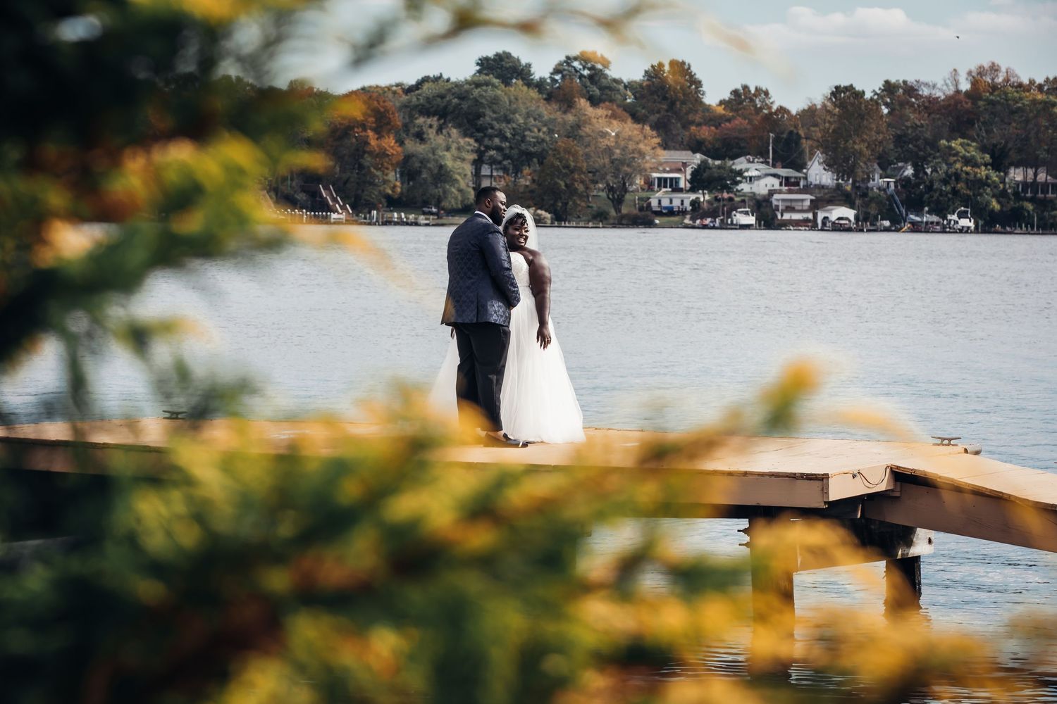 A bride and groom stand together on a wooden dock overlooking a serene lake framed by autumn foliage.