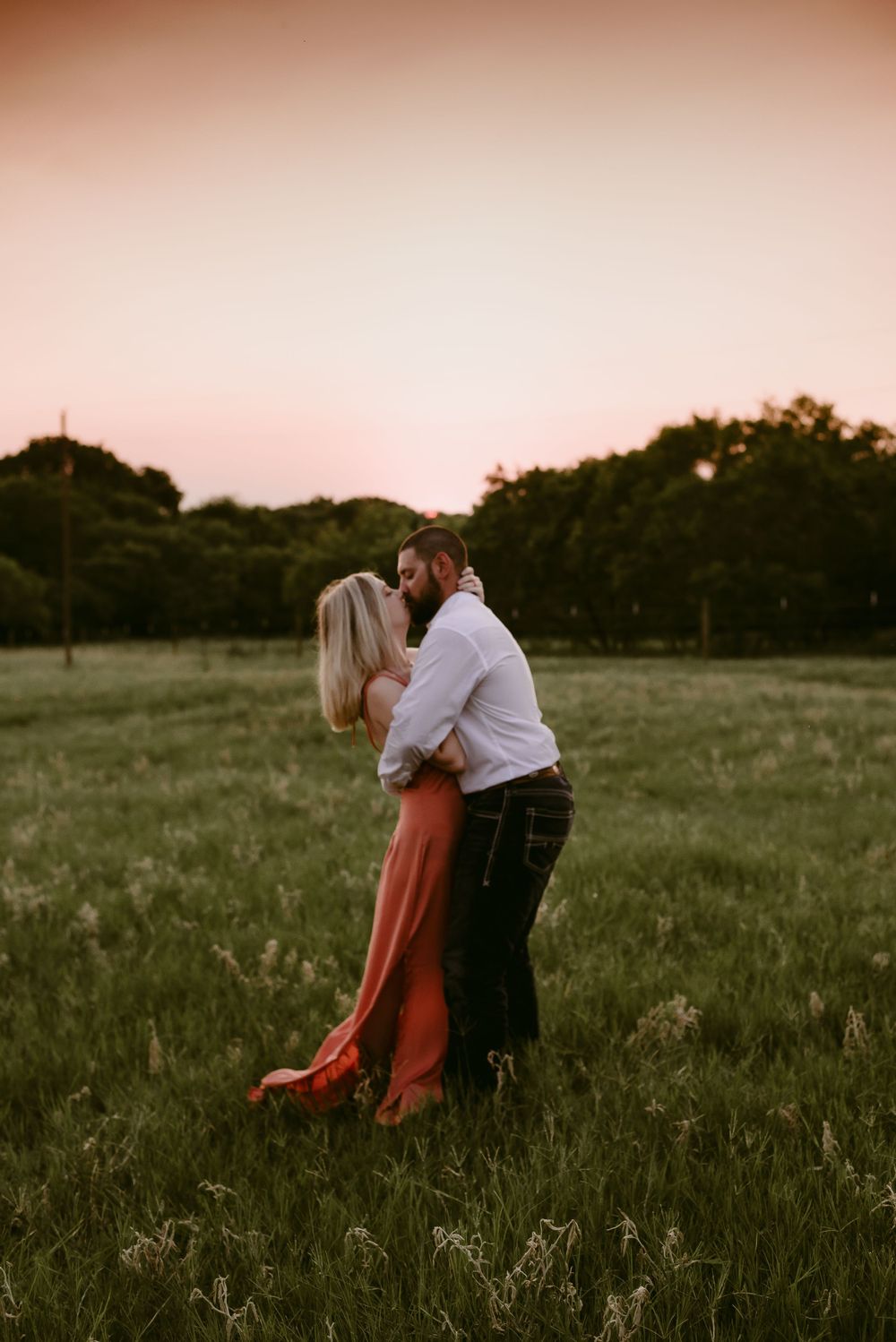 Couple in romantic embrace standing in tall grass during sunset.