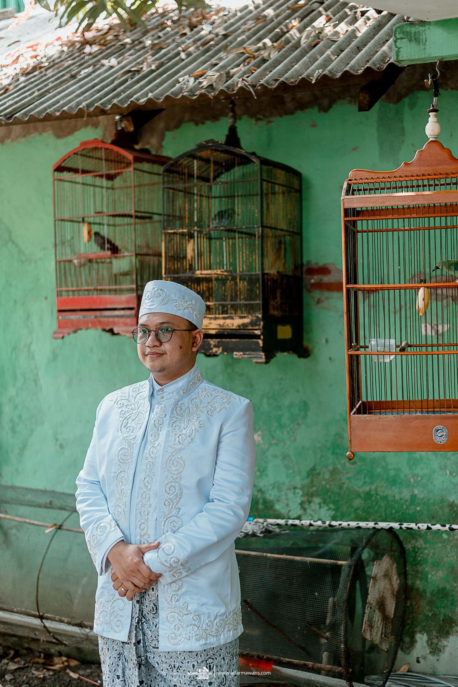 Traditional groom in light blue formal attire stands in front of green wall with decorative bird cages.
