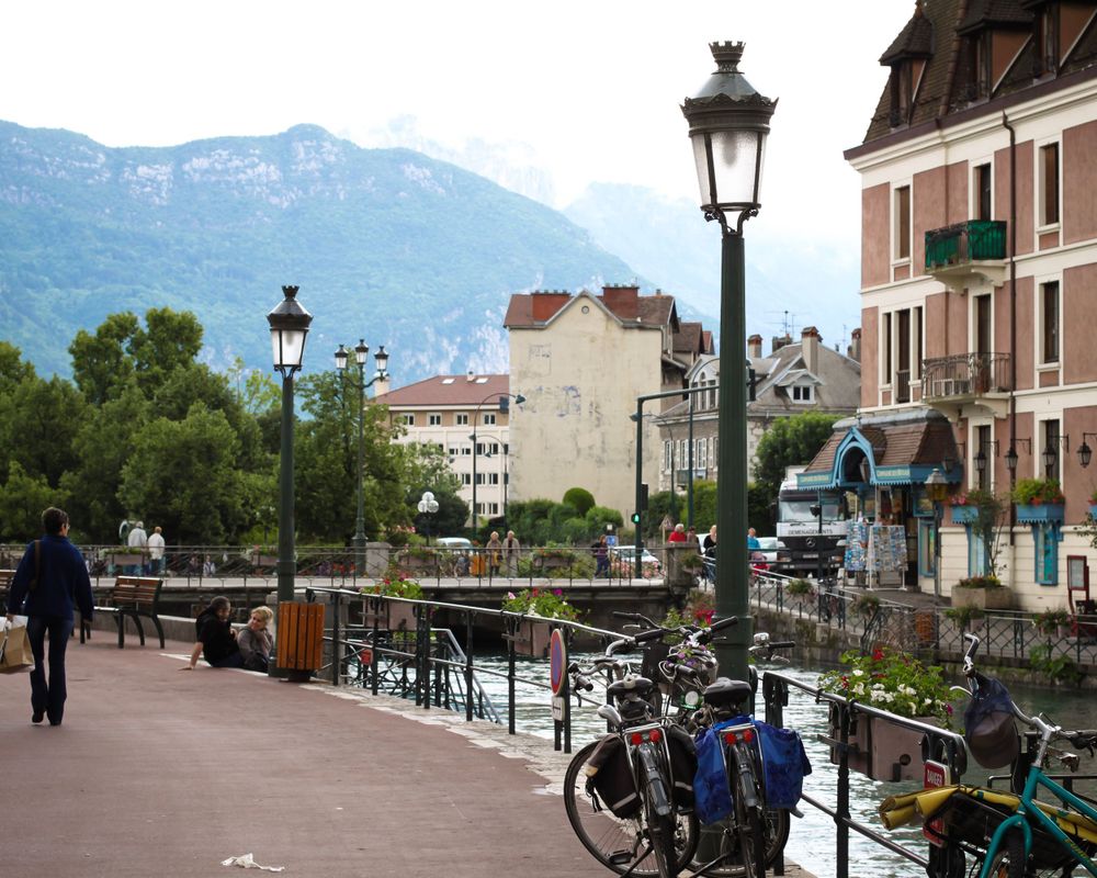 Scenic walkway along a canal in Annecy, France with vintage lampposts and mountains in the background.