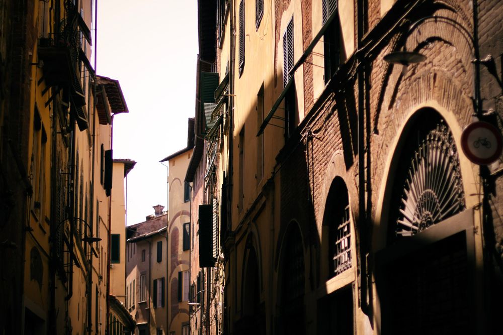 Narrow medieval street in Italy with tall buildings and arched architecture creating dramatic shadows.