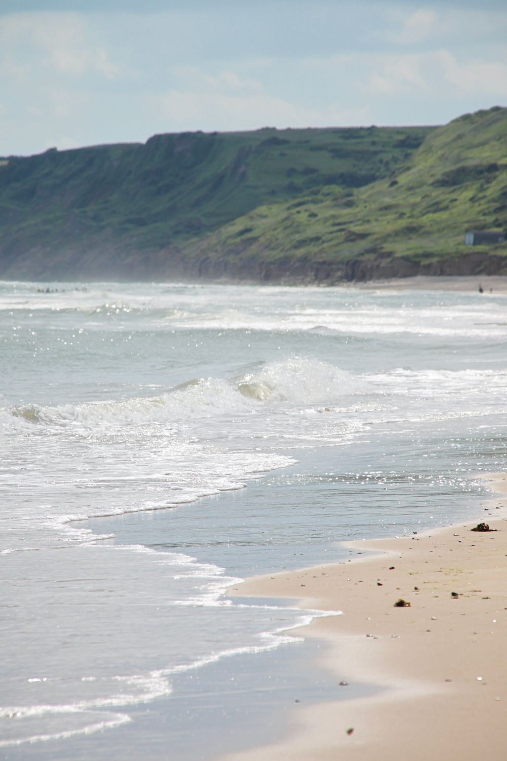 Coastal landscape with rolling green cliffs and gentle waves breaking on a sandy beach.