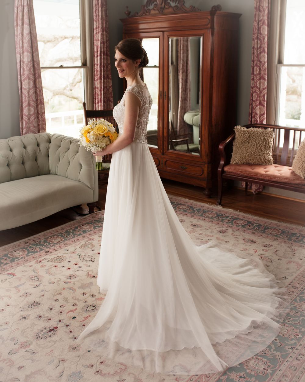 Bride in flowing white gown holding yellow bouquet poses in vintage-styled room.